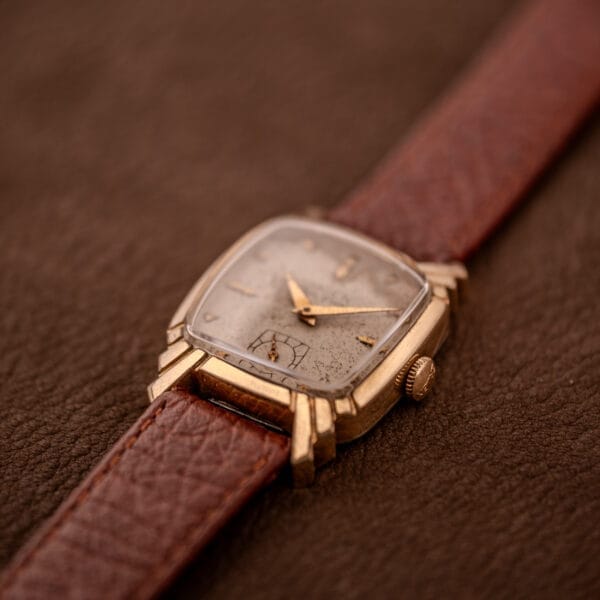 A Gruen Veri-Thin Ref. 415-810 with Art Deco lugs, a brown leather strap, and a gold square case rests on a brown textured surface.