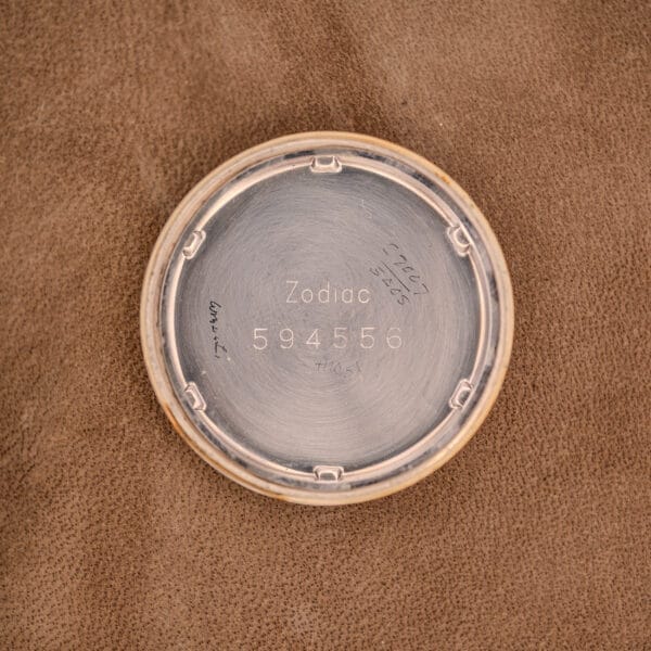 The inside caseback of a Zodiac Hermetic Black Dial Automatic Caliber 1624 Gold Filled Case watch, engraved with Zodiac 594556 and handwritten notes, is shown resting on brown fabric.