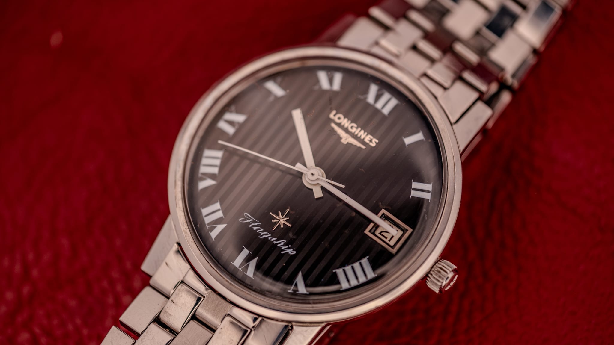 A silver Longines wristwatch with a black dial and Roman numerals, featuring its Serial Number engraved on the back, rests on a red textured surface.