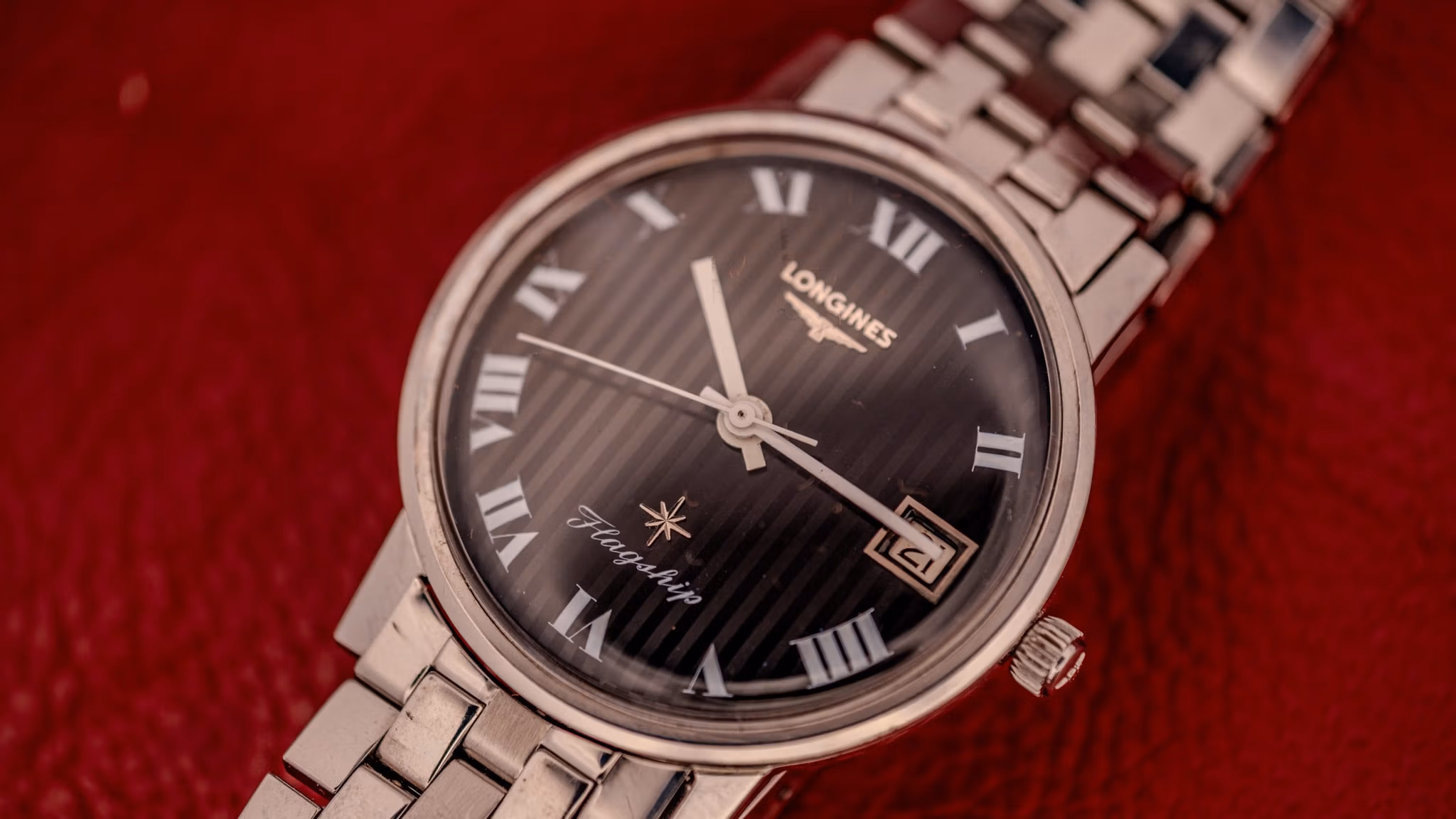 A silver Longines wristwatch with a black dial and Roman numerals, featuring its Serial Number engraved on the back, rests on a red textured surface.