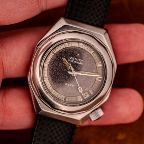 A hand holding a Zenith Automatic Defy NA.50.012.2 in stainless steel Cal. 2562 PC, featuring a textured black dial and black strap.