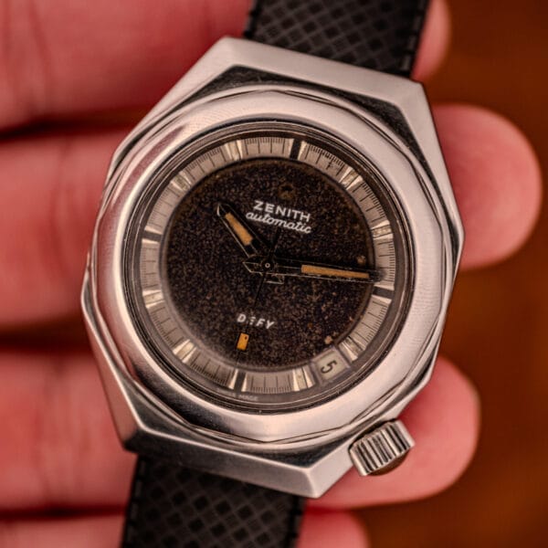 A hand holds a Zenith Automatic Defy NA.50.012.2 in Stainless Steel Cal. 2562 PC, featuring a black textured dial and a date display at 4 o'clock.