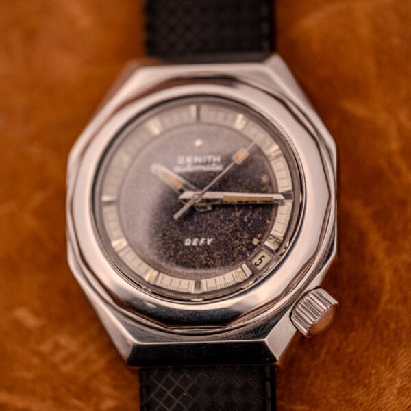 A Zenith Automatic Defy NA.50.012.2 in stainless steel (Cal. 2562 PC) with a textured dark dial and black strap rests on a brown surface, highlighting its classic design.