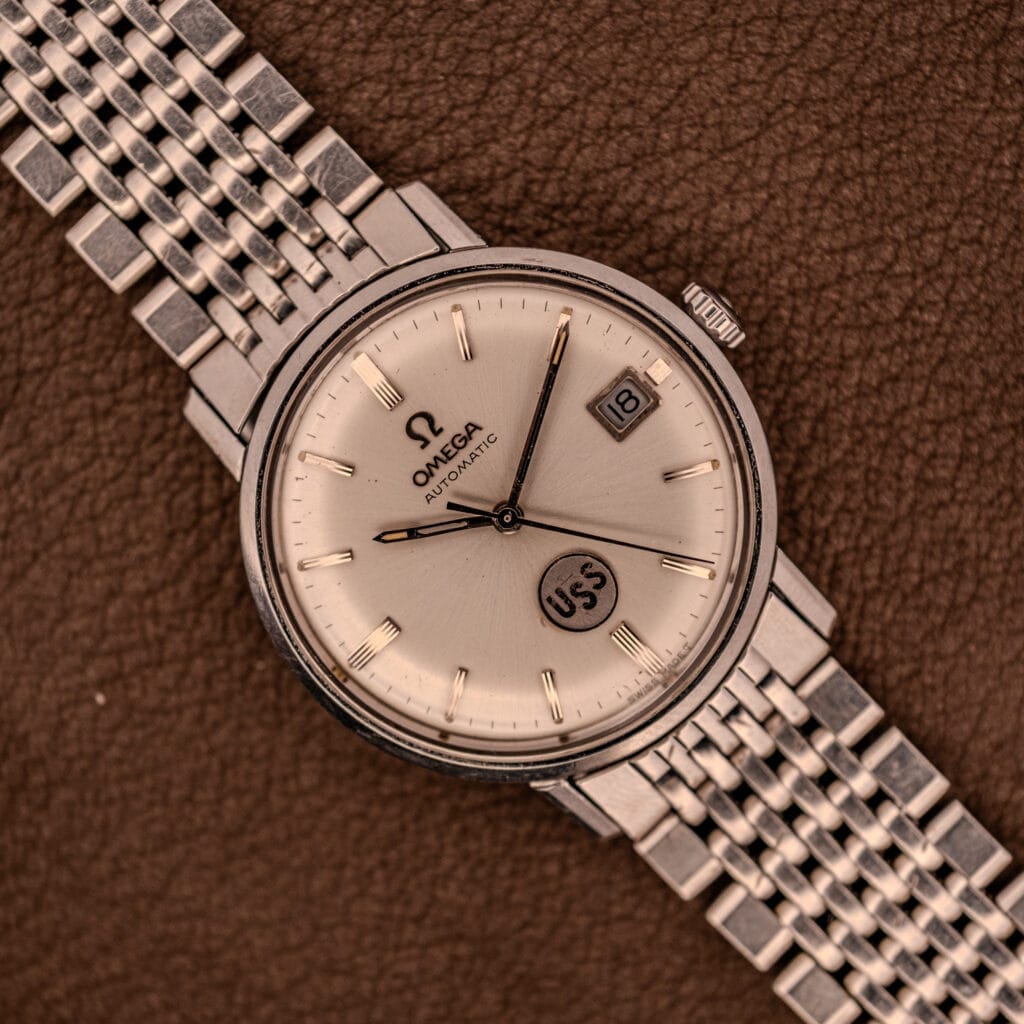 An Omega Automatic Ref. 166.020 for U.S. Steel Presentation Watch, featuring a silver metal band, date display, and USS dial detail, is showcased on brown leather.