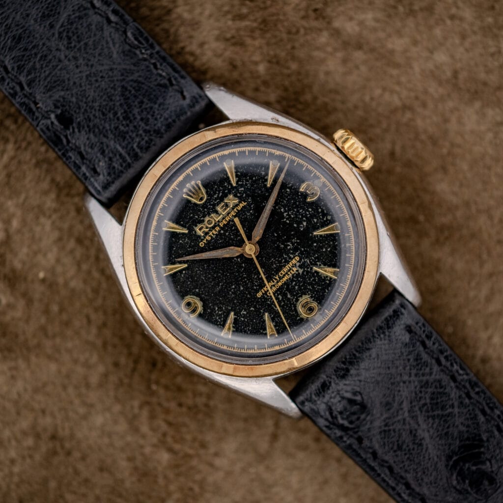 The Rolex Oyster Perpetual "Big Bubble Back" Ref. 6107 Black Dial rests elegantly on a brown textured surface, showcasing its timeless design and classic appeal.