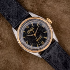 The Rolex Oyster Perpetual "Big Bubble Back" Ref. 6107 Black Dial rests elegantly on a brown textured surface, showcasing its timeless design and classic appeal.