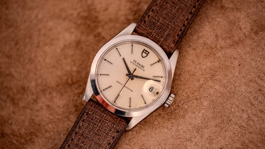 A classic Tudor wristwatch with a brown strap rests elegantly on a tan textured surface.