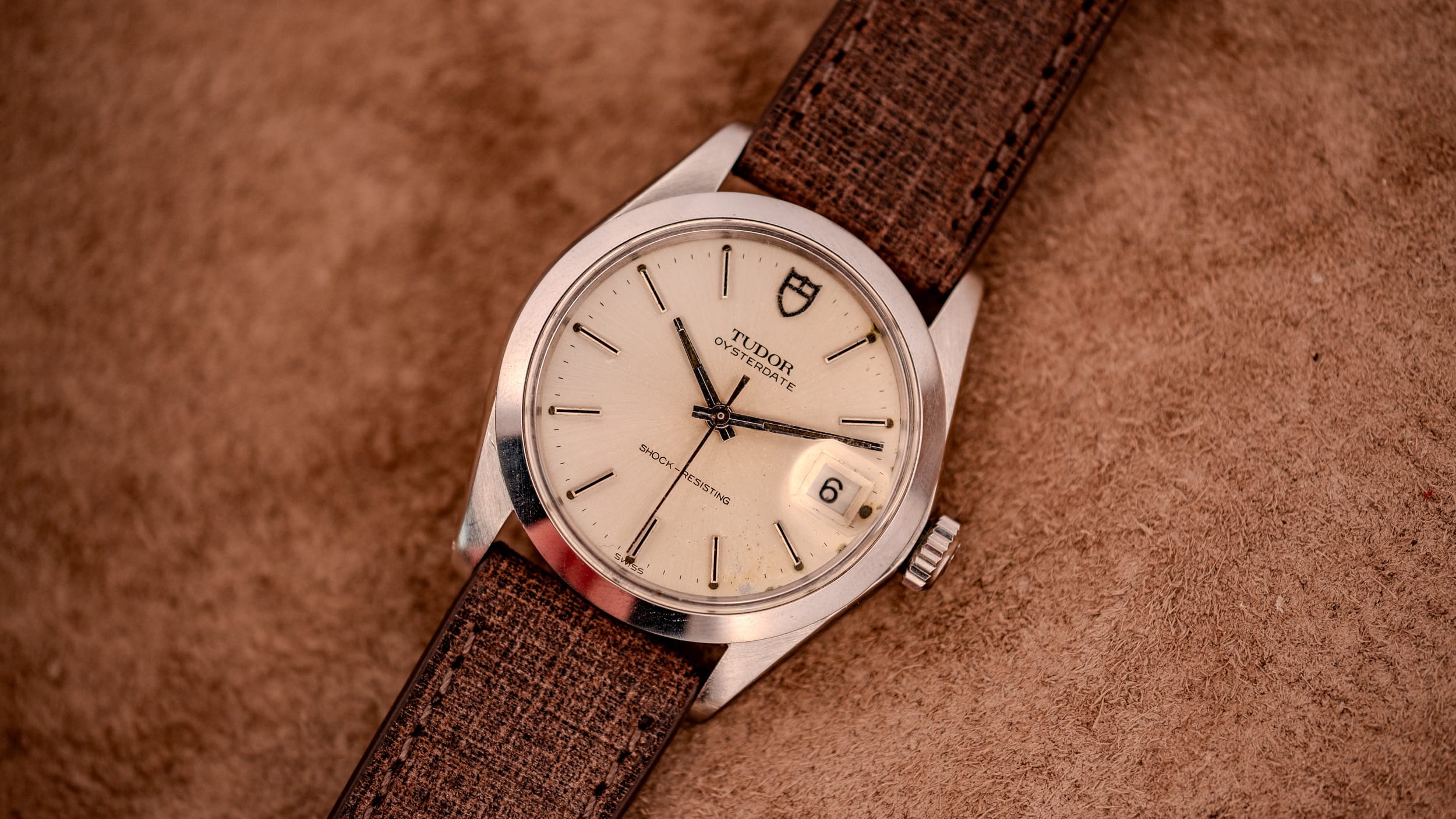 A classic Tudor wristwatch with a brown strap rests elegantly on a tan textured surface.