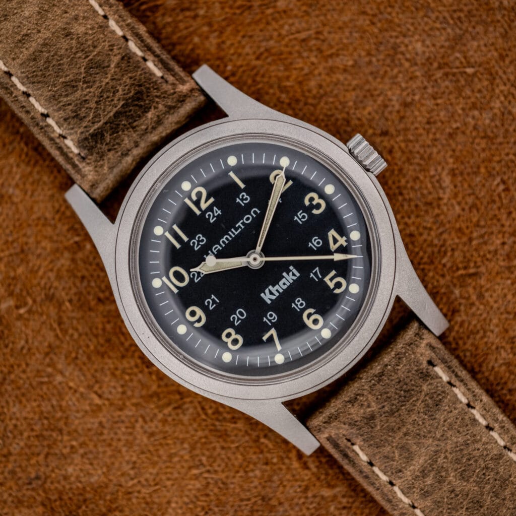 The Hamilton Khaki Ref. 9415A features a black dial and manual wind movement, paired with a brown leather strap, resting on a textured surface to highlight its classic stainless steel design.