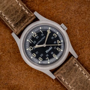 The Hamilton Khaki Ref. 9415A features a black dial and manual wind movement, paired with a brown leather strap, resting on a textured surface to highlight its classic stainless steel design.