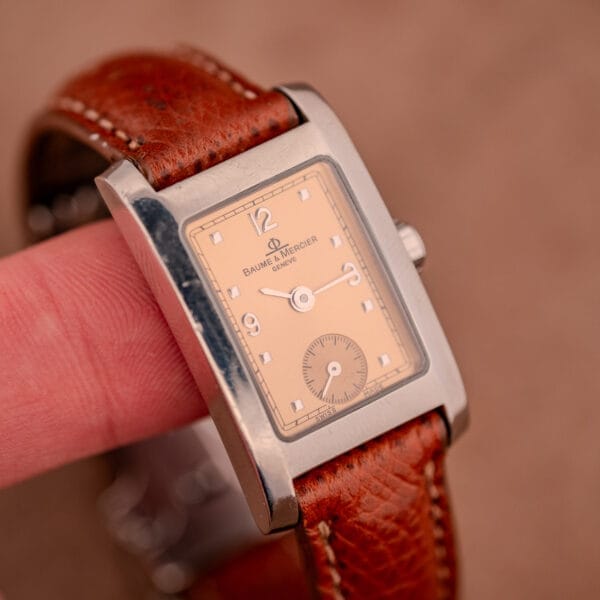A Baume Mercier Hampton MV045139 with a copper dial and stainless steel case is held between fingers, highlighting its refined design.