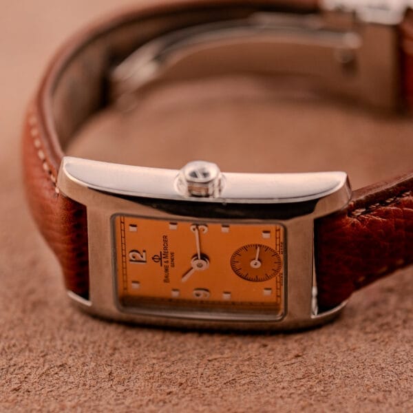 The Baume Mercier Hampton MV045139 wristwatch, featuring a copper dial, stainless steel case, and brown leather strap, rests on a textured brown surface.