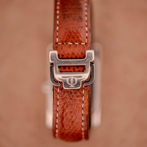 Close-up of a brown leather watch strap with a silver buckle, set against a tan background, evoking the refined elegance of the Baume Mercier Hampton MV045139 Copper Dial Stainless Steel.