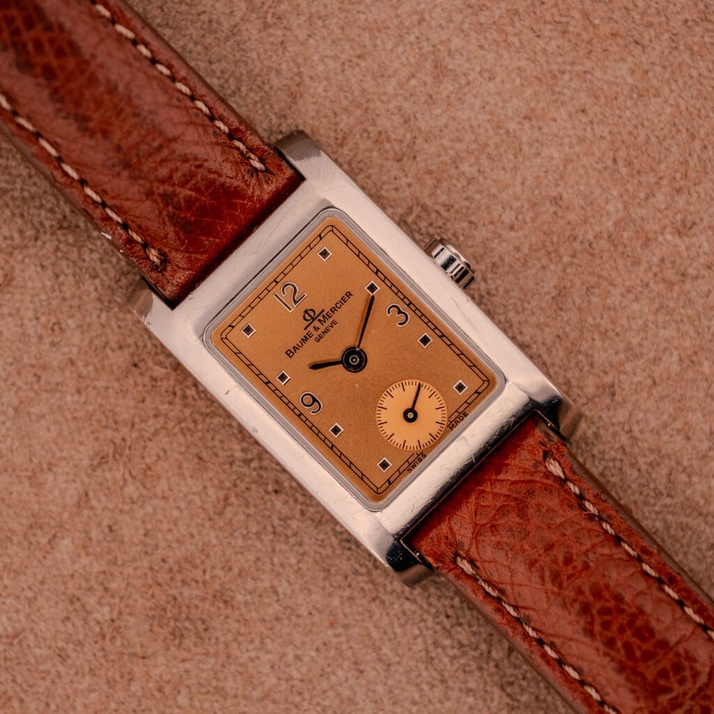 Baume Mercier Hampton MV045139 watch with a copper dial and stainless steel case, displayed on a tan textured surface.
