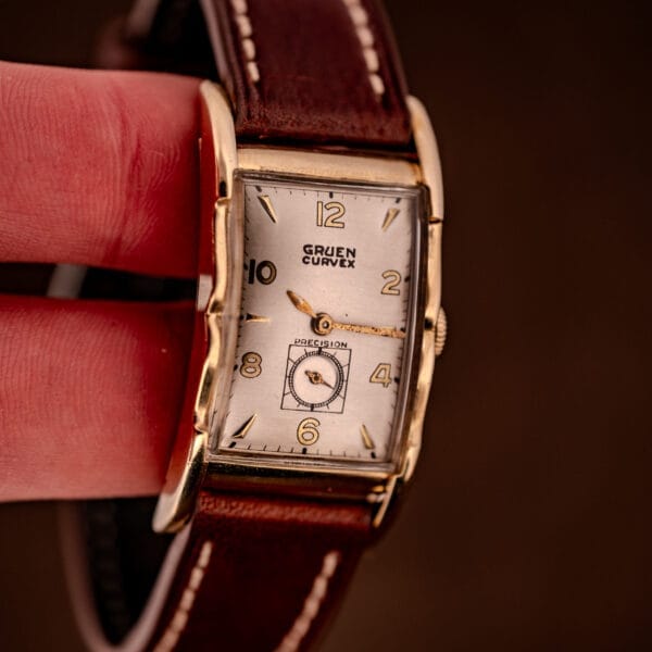 A hand holding the Gruen Curvex Precision Ref. 370-600 wristwatch, featuring a rectangular 10k gold filled case and a brown leather strap.