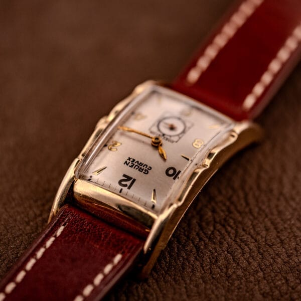 A Gruen Curvex Precision Ref. 370-600 wristwatch with a 10k gold filled case and brown leather strap rests elegantly on a textured brown surface.