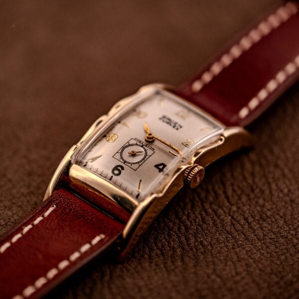 A Gruen Curvex Precision Ref. 370-600 wristwatch with a 10k gold filled case and brown leather strap rests on a textured brown surface.