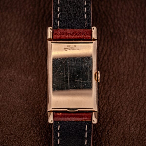Gruen Curvex Precision Ref. 370-600 wristwatch with a rectangular 10k gold filled case and black leather strap, displayed on a brown textured background.