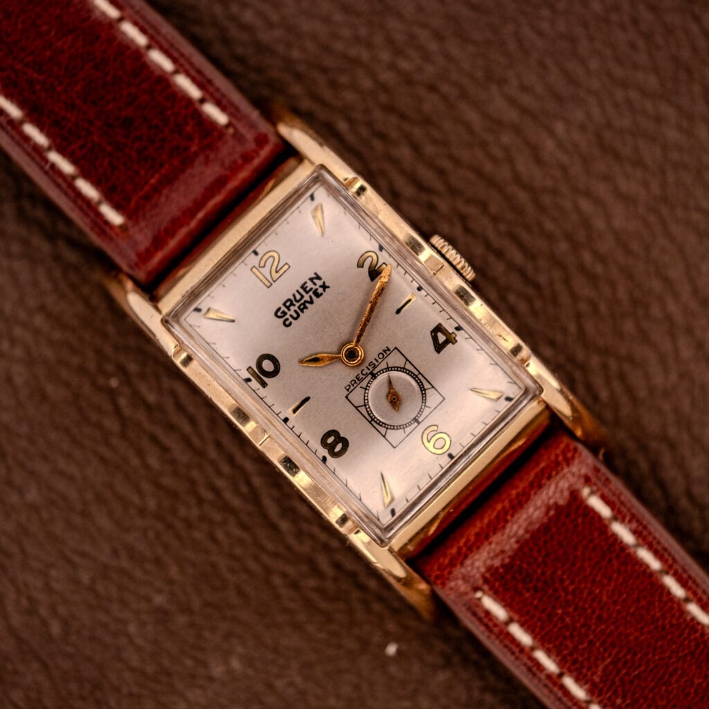 A Gruen Curvex Precision Ref. 370-600 wristwatch with a 10k gold filled case and brown leather strap rests on a brown textured surface.