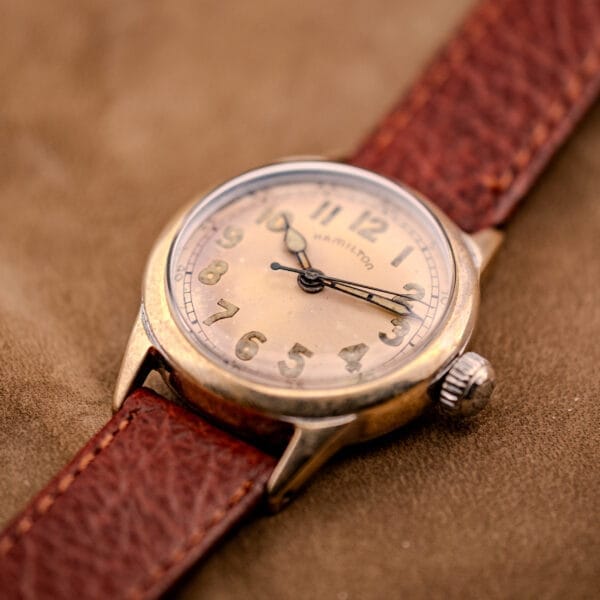 Close-up of a Hamilton WWII Era U.S. Military Cal. 987S Hacking Seconds wristwatch with a brown leather strap, displayed on a tan surface.