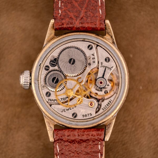 Close-up of a vintage Hamilton WWII Era U.S. Military Cal. 987S Hacking Seconds watch with exposed gears and a brown leather strap, set against a brown background.