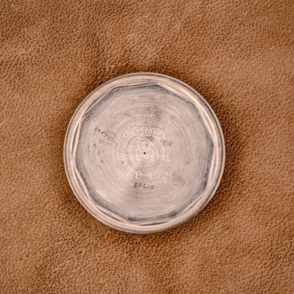 A round metal object with engraved text, resembling the case back of a Hamilton WWII Era U.S. Military Cal. 987S Hacking Seconds watch, rests on a brown textured surface.