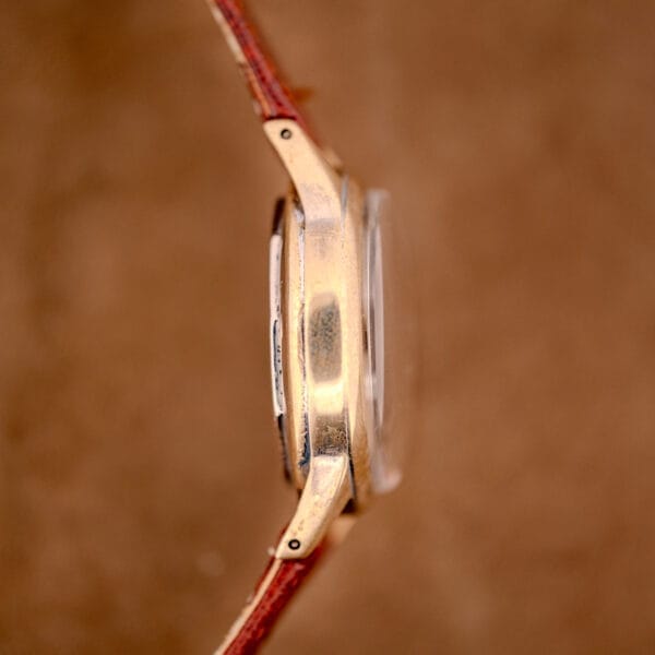 Side view of a Hamilton WWII Era U.S. Military Cal. 987S Hacking Seconds watch with a gold case and brown leather strap, set against a brown background.