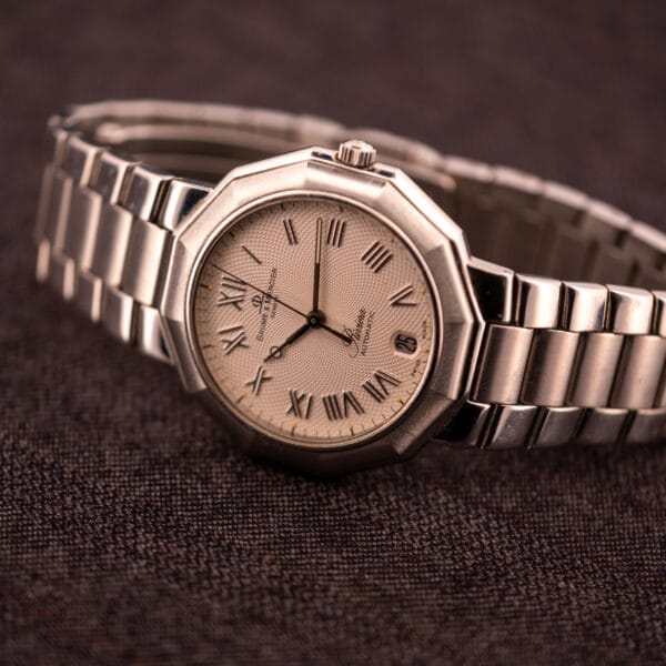 A Baume Mercier Riviera Automatic MV045165 with a Guilloché dial and Roman numeral markers rests on textured dark brown fabric.