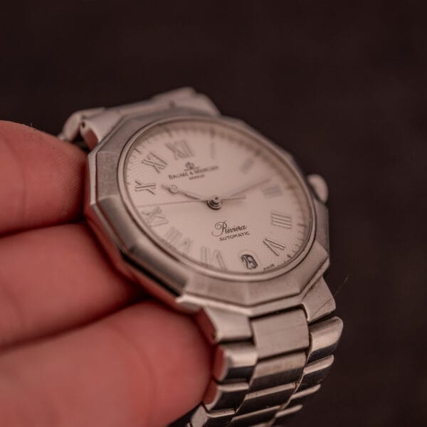 A hand holds a Baume Mercier Riviera Automatic MV045165 wristwatch with a silver case, white guilloché dial, and Roman numerals.