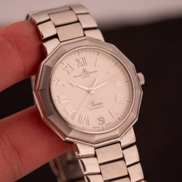 A hand holding a Baume Mercier Riviera Automatic MV045165 wristwatch with a silver case and white guilloché dial.