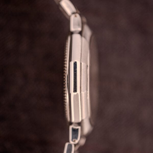 Side view of the Baume Mercier Riviera Automatic MV045165 with a silver metal case and bracelet and guilloché dial, against a blurred brown textured background.