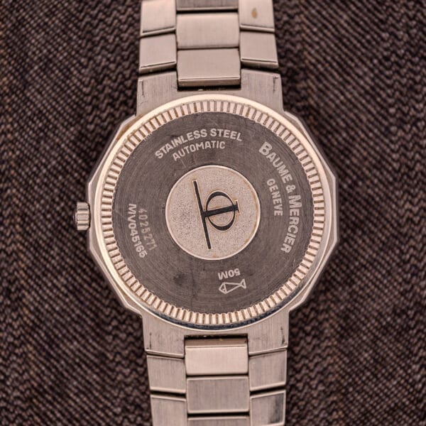 The Baume Mercier Riviera Automatic MV045165 watch features its engraved back on a textured dark fabric, showcasing the refined craftsmanship of its Guilloché Dial.