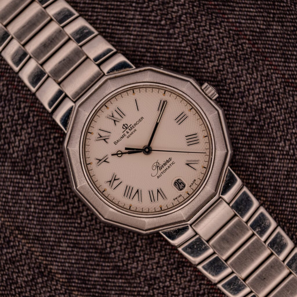A Baume Mercier Riviera Automatic MV045165 wristwatch with a guilloché dial is displayed on a textured brown fabric background.