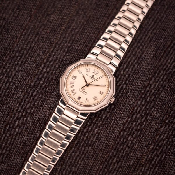 Baume Mercier Riviera Automatic MV045165 wristwatch with a Guilloché dial and Roman numerals on a metallic band, displayed on textured brown fabric.