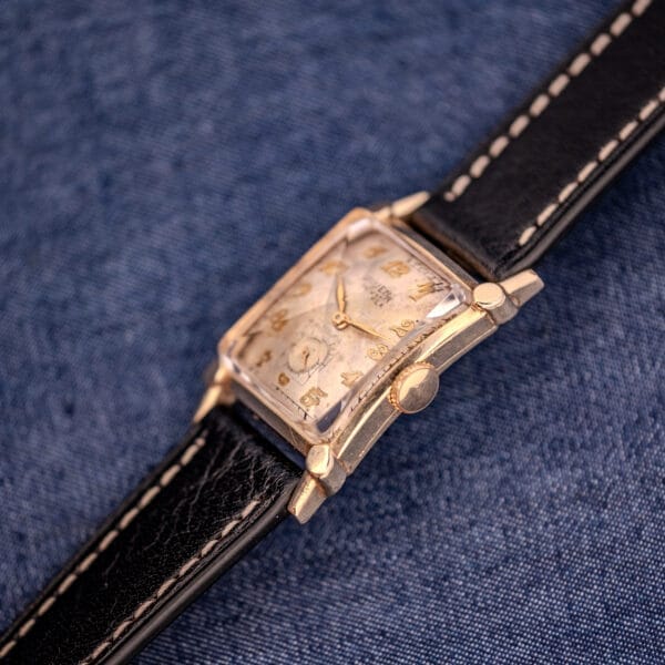 The Gruen Curvex Ref. 370-642 Art Deco "Pyramid" Crystal wristwatch, featuring a gold case and black leather strap on blue denim fabric, exudes vintage elegance and timeless style.