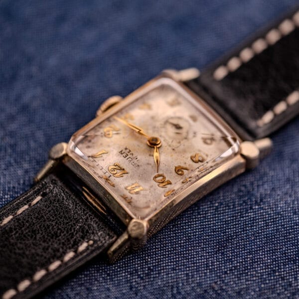 A Gruen Curvex Ref. 370-642 Art Deco "Pyramid" Crystal wristwatch with a worn dial and black leather strap rests on blue fabric, highlighting its vintage charm.