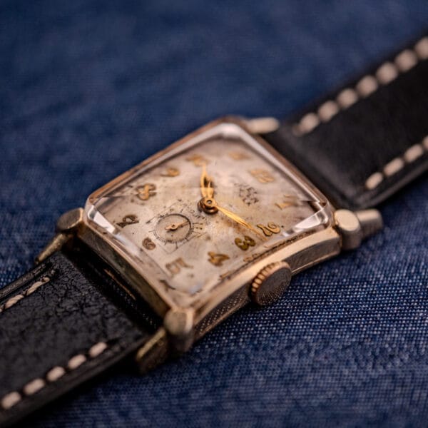 A Gruen Curvex Ref. 370-642 Art Deco "Pyramid" Crystal wristwatch with a worn face and black leather strap rests on blue fabric, highlighting its vintage charm.