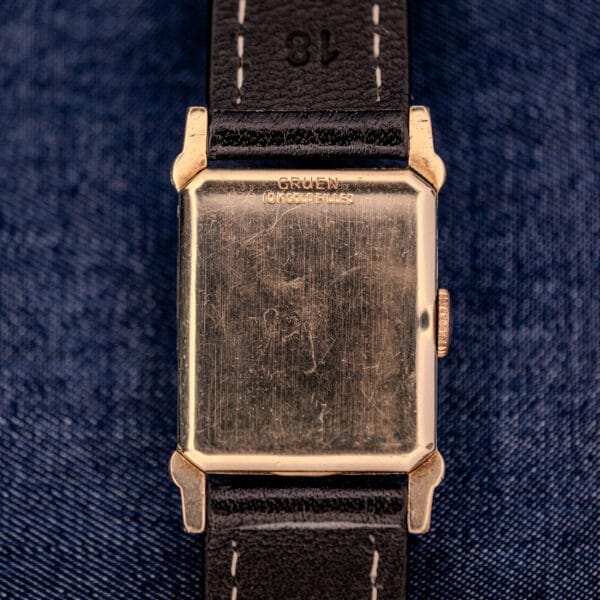 A Gruen Curvex Ref. 370-642 Art Deco "Pyramid" Crystal wristwatch with gold case and black leather strap rests on dark blue denim fabric.