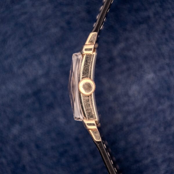 Side view of a Gruen Curvex Ref. 370-642 Art Deco "Pyramid" Crystal gold wristwatch with a black leather strap, set against a blue denim background.