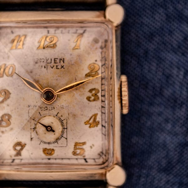 Close-up of a Gruen Curvex Ref. 370-642 Art Deco "Pyramid" Crystal wristwatch with a square face, gold numbers, and unique pyramid crystal detail on a blue fabric background.