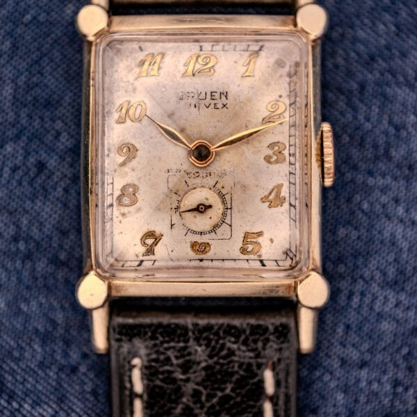 A Gruen Curvex Ref. 370-642 Art Deco "Pyramid" Crystal watch with a vintage square face and black leather strap rests on blue fabric.