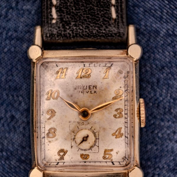 Close-up of a vintage Gruen Curvex Ref. 370-642 Art Deco "Pyramid" Crystal wristwatch with a worn gold face and black leather strap on blue fabric, highlighting its subtle Art Deco elegance.
