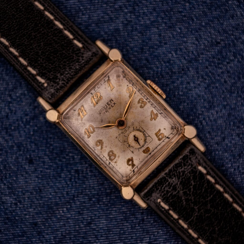 A Gruen Curvex Ref. 370-642 Art Deco "Pyramid" Crystal wristwatch with a rectangular face and black leather strap rests elegantly on blue denim fabric.