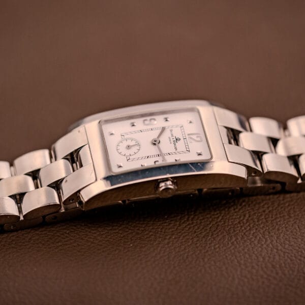 A Baume Mercier Hampton XL MV045063 watch with a white dial and stainless steel band is placed on a brown surface.