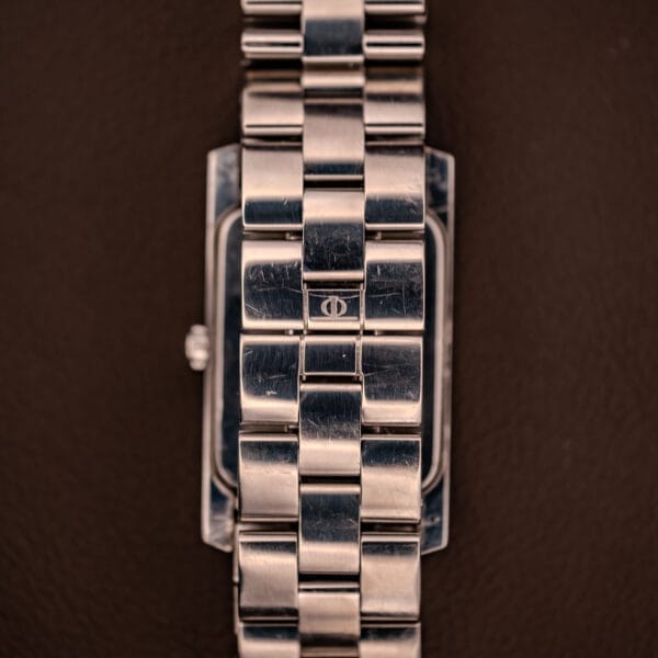 Close-up of a Baume Mercier Hampton XL MV045063 watch band in stainless steel, featuring a white dial and small CD logo, set against a brown background.