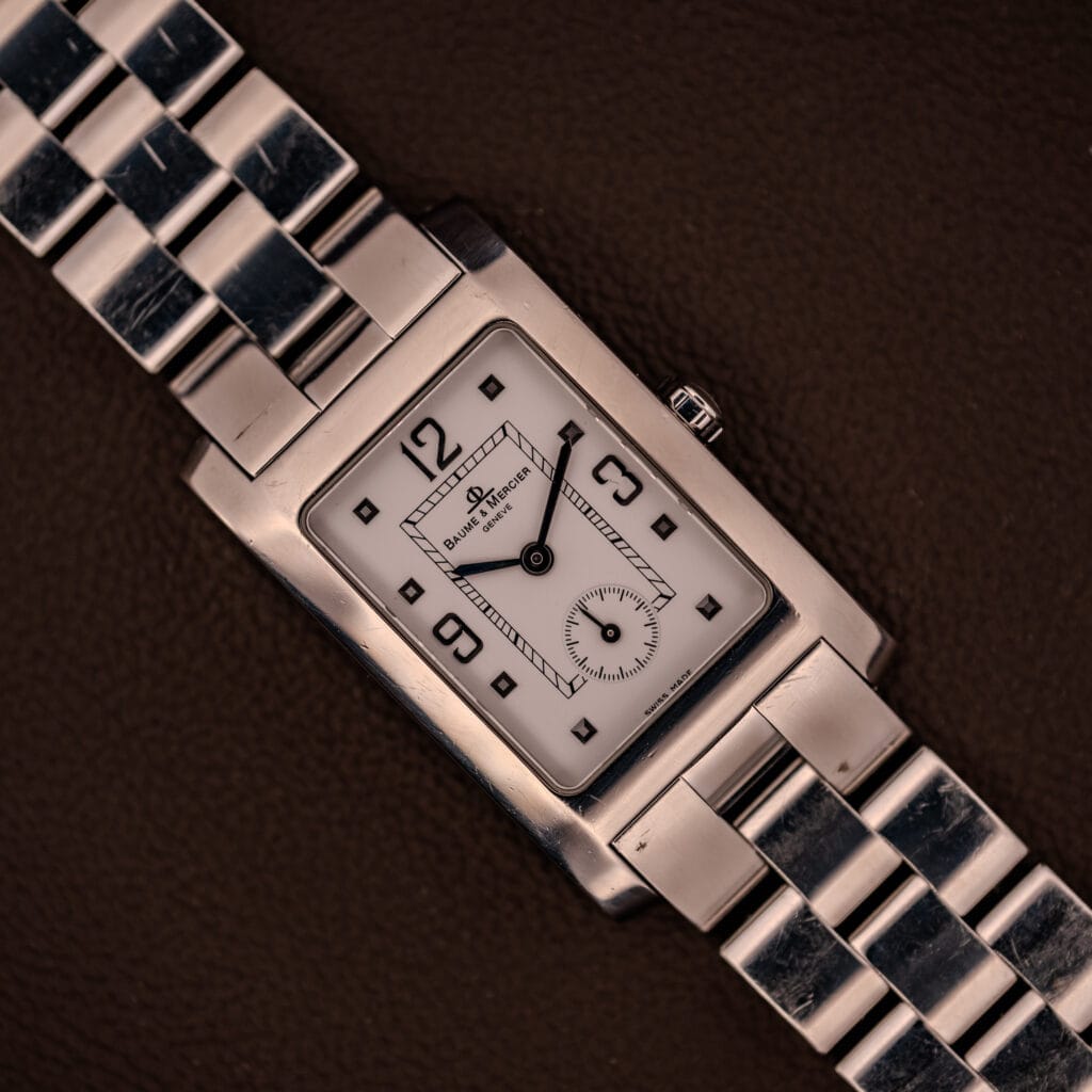 The Baume Mercier Hampton XL MV045063 wristwatch features a white dial and stainless steel band, showcased against a brown background.