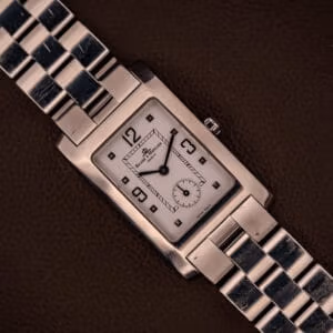 The Baume Mercier Hampton XL MV045063 wristwatch features a white dial and stainless steel band, showcased against a brown background.