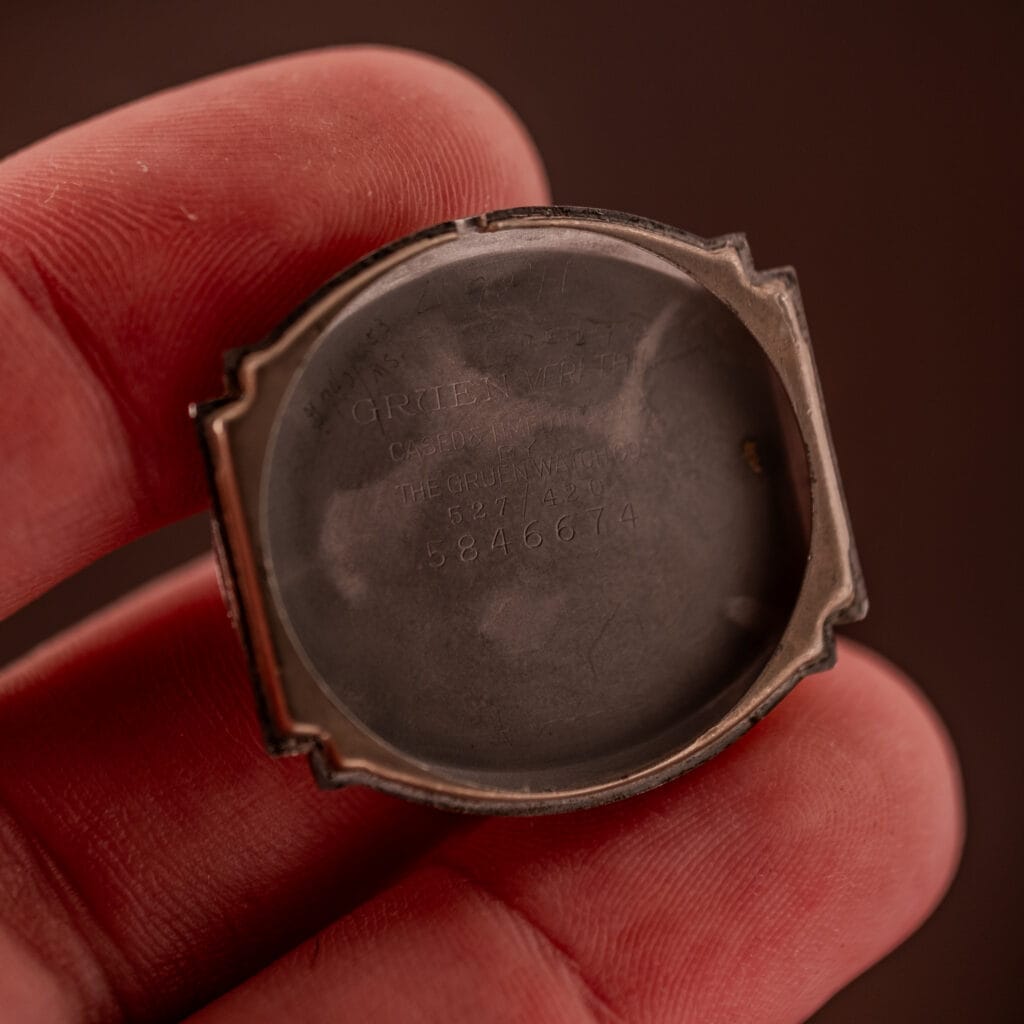 A close-up of a hand holding an empty Gruen Veri-Thin Ref. 527/420 Art Deco Hooded Lugs watch case, highlighting engraved text inside the back cover and classic Art Deco details.