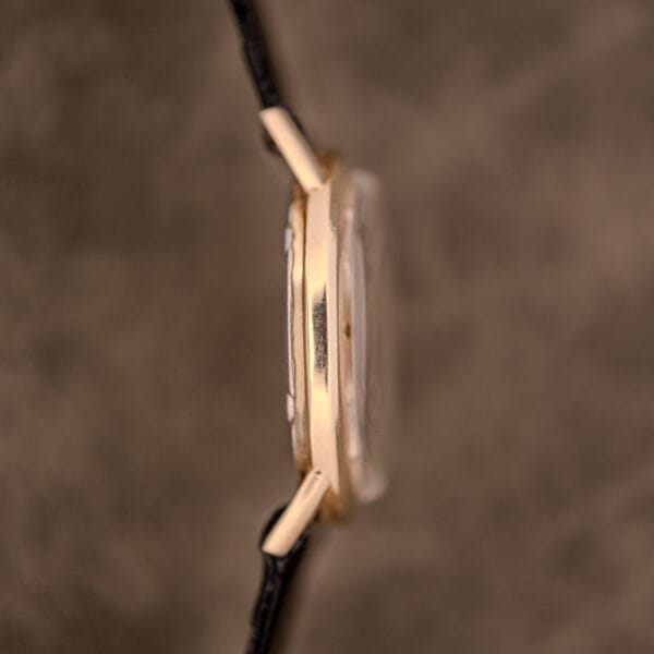 Side view of an Omega Automatic Ref. LU 6304 "HF" Logo Dial wristwatch with a 10k GF bezel and black leather strap, set against a soft brown background.