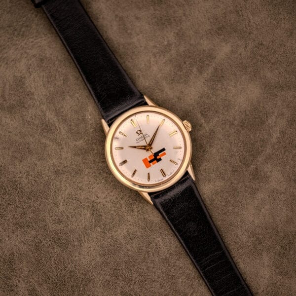 An Omega Automatic Ref. LU 6304 "HF" Logo Dial wristwatch with a 10k GF bezel and black leather strap rests on a brown textured surface.